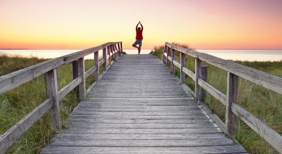 Eine Person steht auf einem Holzsteg Richtung Nordsee und macht eine Yoga-Figur, © AdobeStock_524019202