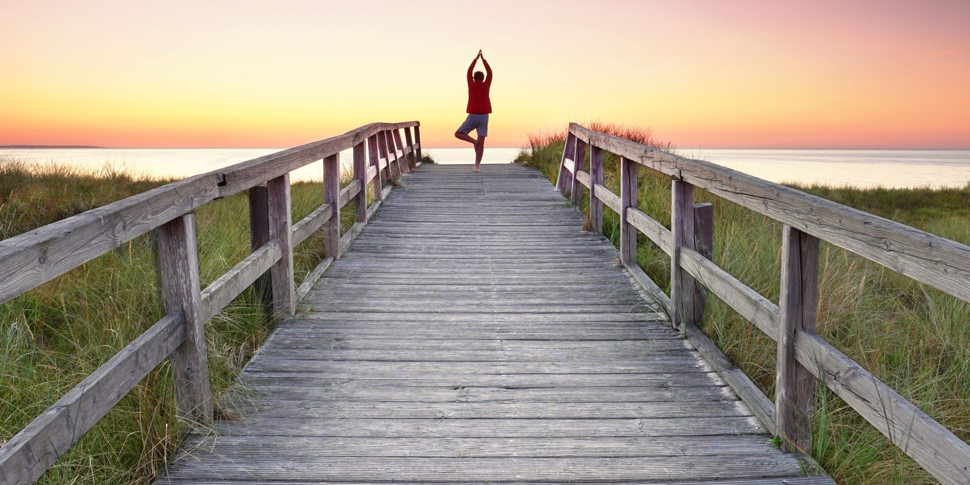 Eine Person steht auf einem Holzsteg Richtung Nordsee und macht eine Yoga-Figur, © AdobeStock_524019202