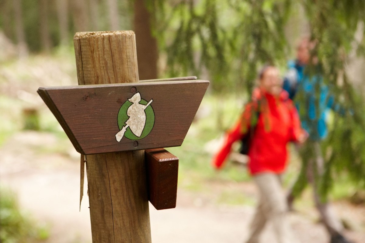 Im Vordergrund das Wegeschild der Wandertour, im Hintergrund ein wanderndes Pärchen., © Harzer Tourismusverband / M.  Gloger